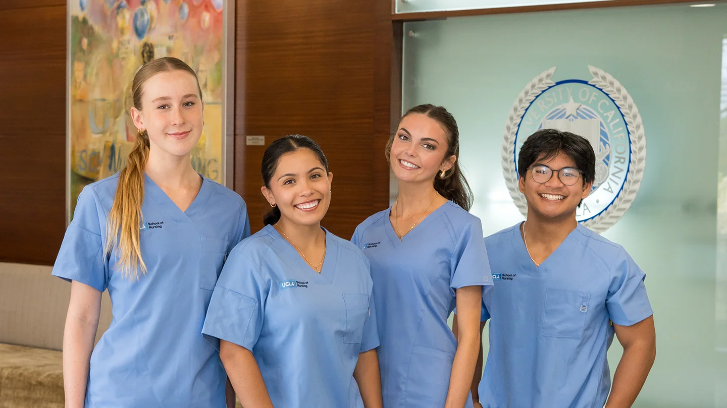 4 nursing students wearing blue scrubs smiling for a photograph in the Factor Building lobby with the new Joe C. Wen School of Nursing sign visible.