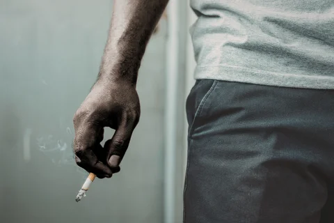 Close-up of a person’s hand holding a lit cigarette with smoke rising, next to their hip, wearing a light gray shirt and dark pants against a neutral background.