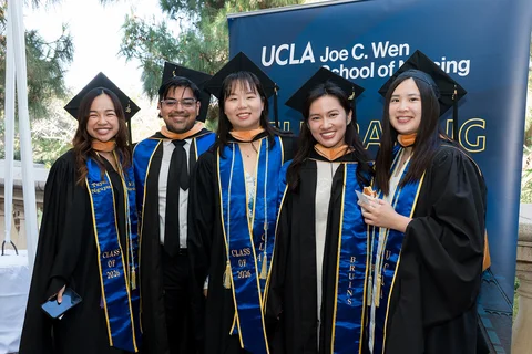 5 UCLA nursing students wearing their graduation caps and gowns standing in front of a UCLA Joe C. Wen School of Nursing sign.