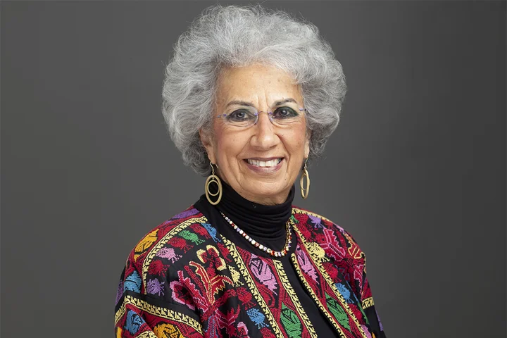 Afaf Meilis is shown in a studio portrait against a gray background, wearing glasses, hoop earrings, a beaded necklace, and a multicolored patterned jacket over a dark top. 