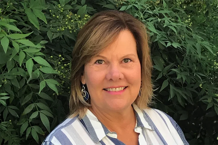Karen Nelson wearing a white and blue stripped shirt standing in front of greenery. 