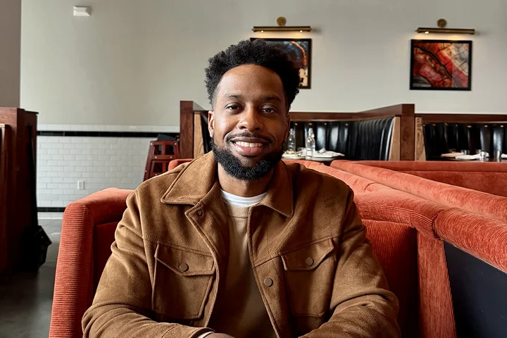Marsellis Irving sits at a restaurant booth with hands resting on the table, wearing a brown jacket over a light-colored shirt.