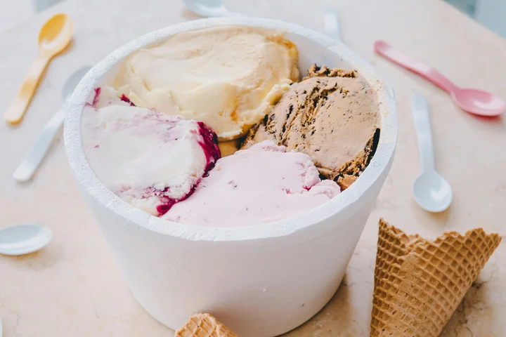 Tub of assorted ice cream flavors with waffle cones and colorful spoons on a table.