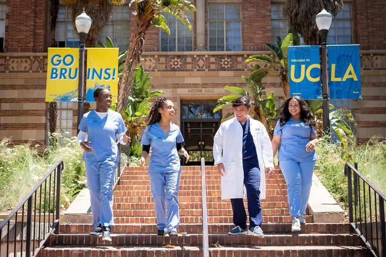 Five people wearing light blue medical scrubs and one white lab coat walk down brick steps on a university campus. Banners on lampposts read “GO BRUINS!” and “WELCOME TO UCLA."