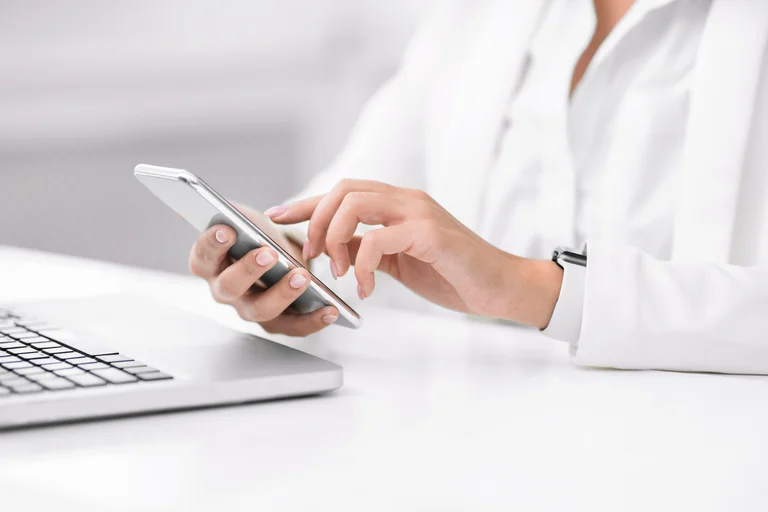 Close-up of a person’s hands using a smartphone on a white desk, with an open laptop nearby; the person wears a white long-sleeve shirt and a smartwatch, in a bright, minimal workspace.