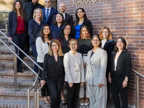 Fifteen DNP students, class of 2026, standing staggered in rows outside on the front steps of the School of Nursing building. Students are dressed in professional clothing and are smiling.
