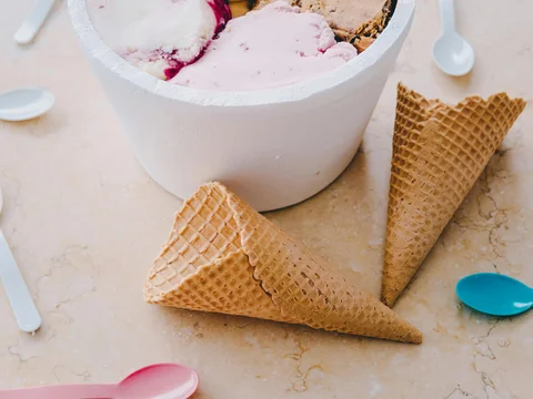 Tub of assorted ice cream flavors with waffle cones and colorful spoons on a table.