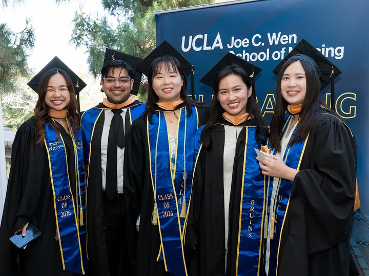 5 UCLA nursing students wearing their graduation caps and gowns standing in front of a UCLA Joe C. Wen School of Nursing sign.