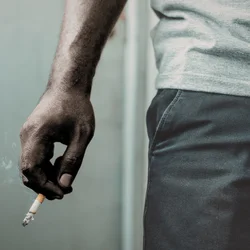 Close-up of a person’s hand holding a lit cigarette with smoke rising, next to their hip, wearing a light gray shirt and dark pants against a neutral background.