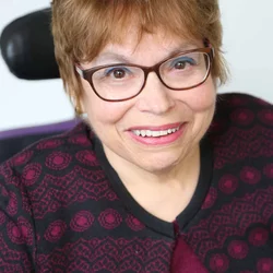 A headshot of Judy Heumann, a cis-gender white woman who is a wheelchair user with short brown hair. She is wearing brown glasses, a maroon and black embroidered cardigan with the top buttoned, and a matching maroon shirt. She is smiling kindly. 