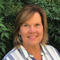 Karen Nelson wearing a white and blue stripped shirt standing in front of greenery. 