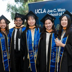 5 UCLA nursing students wearing their graduation caps and gowns standing in front of a UCLA Joe C. Wen School of Nursing sign.