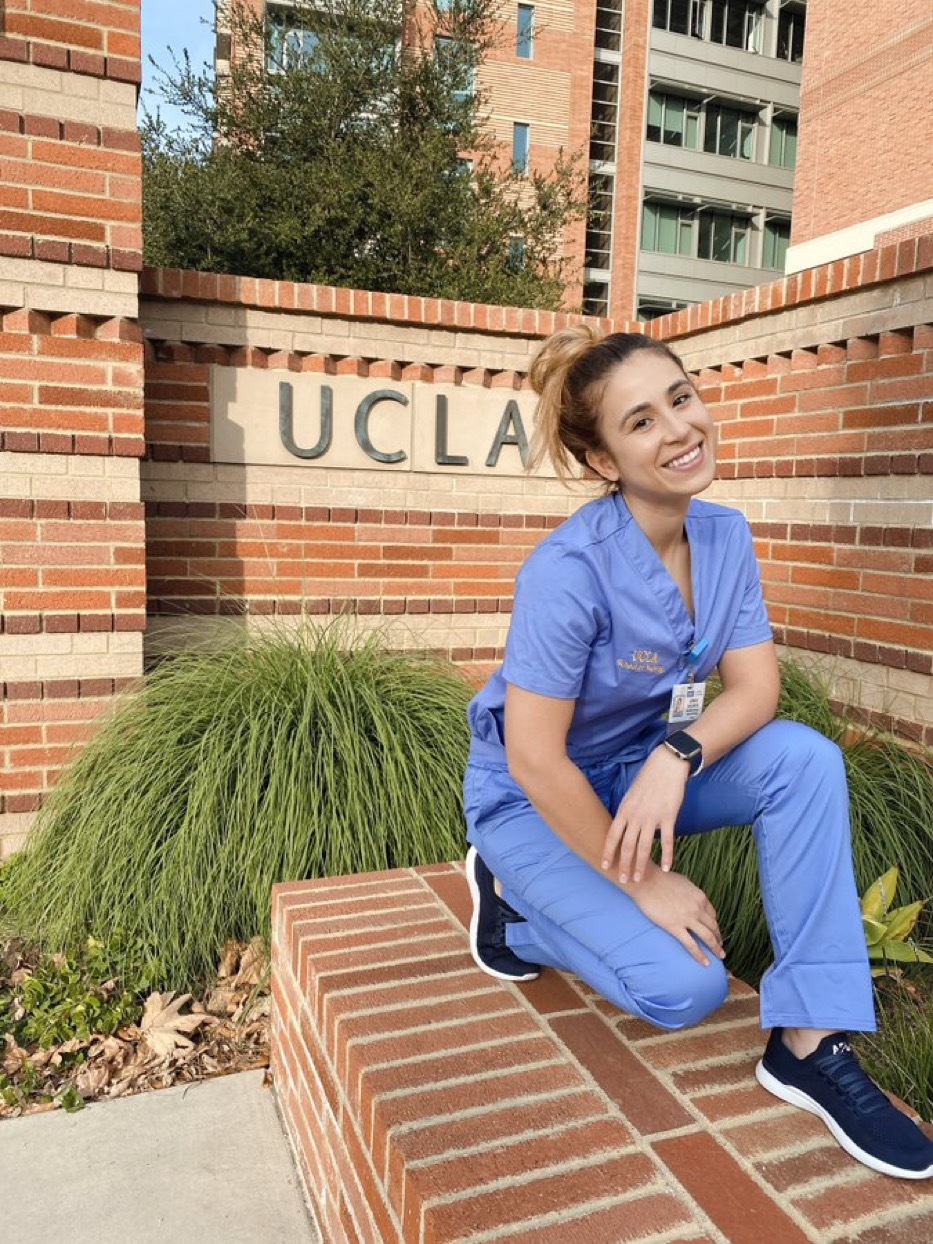 Nursing student in scrubs sitting in front of a UCLA sign on campus